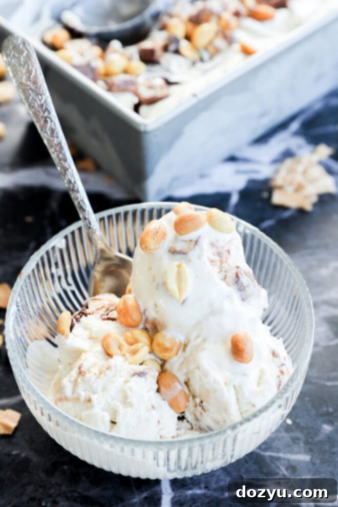 A glass bowl with two scoops of Snickers Ice Cream topped with peanuts, with a silver spoon inside. A loaf pan filled with more creamy ice cream and nuts sits in the background on a dark marble surface.
