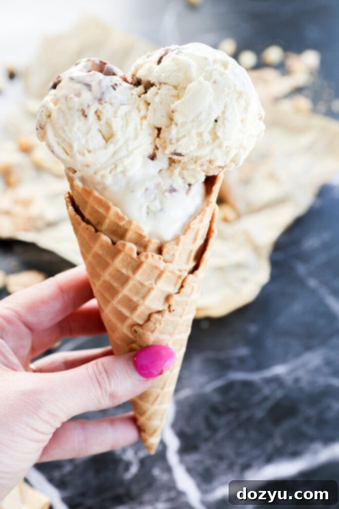 A hand with pink nail polish holds a waffle cone with two scoops of Snickers Ice Cream, possibly with chocolate or nuts, over a dark marble surface and some crumpled parchment paper.