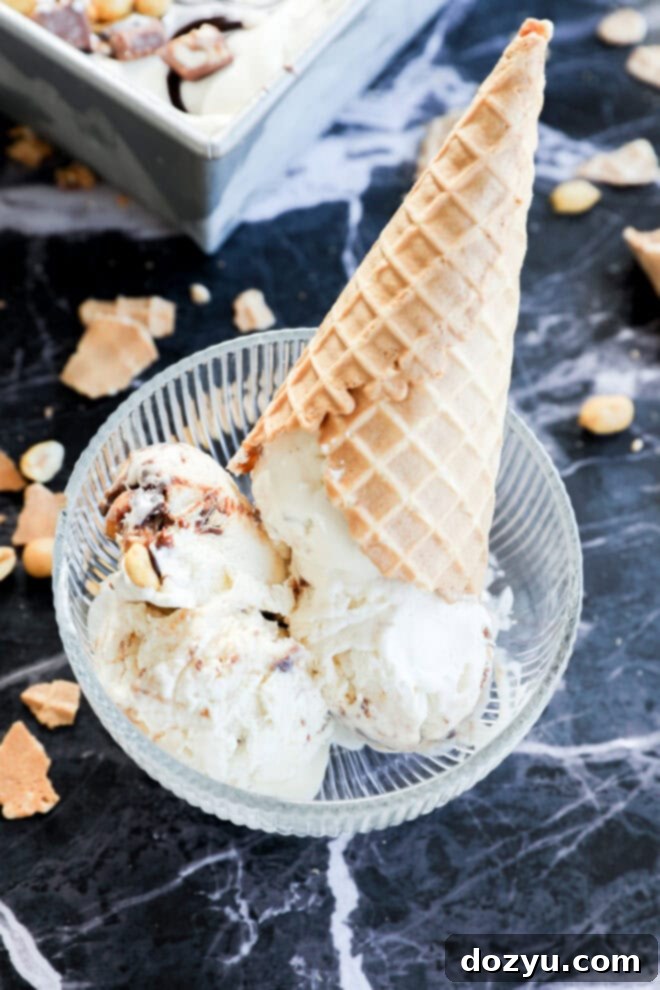 A glass bowl with scoops of Snickers Ice Cream and a waffle cone placed upside down on top, set on a black marble surface with pieces of nuts and cone scattered around.