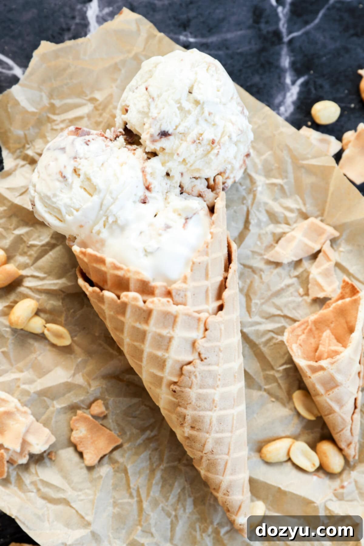 Two scoops of Snickers Ice Cream in a waffle cone rest on crumpled parchment paper, surrounded by scattered peanuts and broken pieces of cone on a dark marble surface.