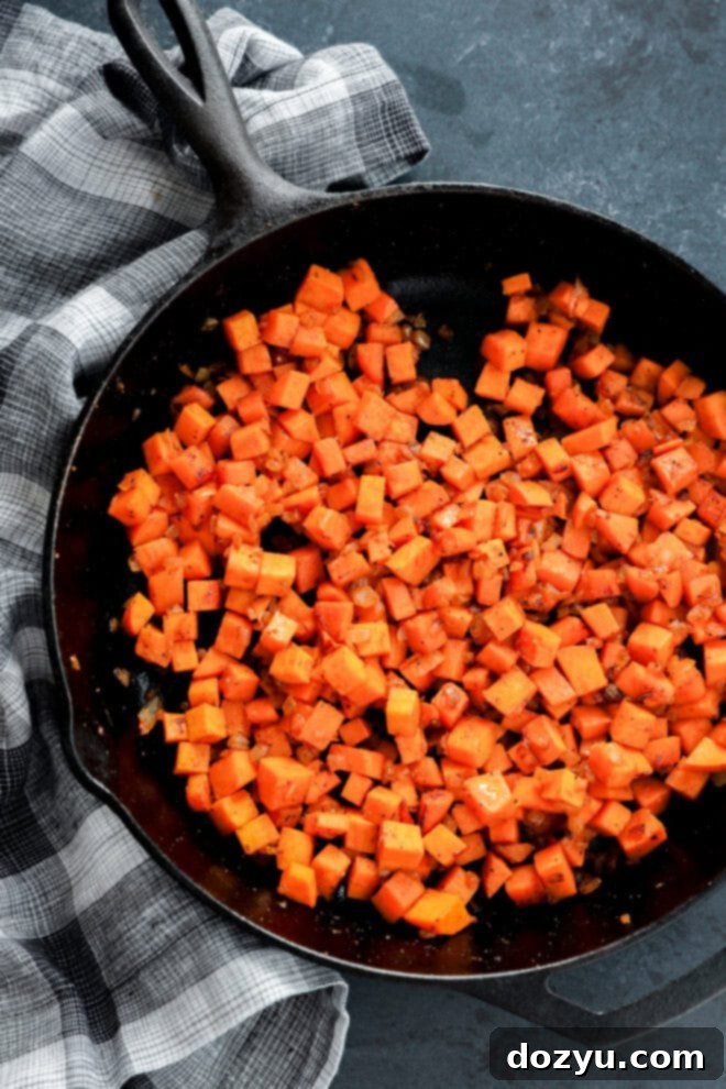 A rustic black cast iron skillet brimming with freshly diced, golden-brown sweet potatoes sautéed to perfection. The potatoes are coated with a medley of aromatic herbs and spices, showcasing a slightly crispy texture and vibrant orange color. The skillet rests on a dark, textured surface, with a classic black and white plaid kitchen towel neatly folded beside it, highlighting the cozy, home-cooked feel of the dish.