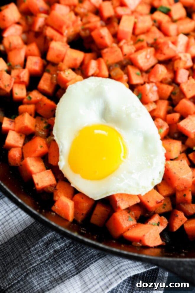 A beautifully cooked fried egg with a perfectly bright yellow, runny yolk rests atop a serving of freshly sautéed sweet potato breakfast potatoes in a black skillet. The dish is artfully garnished with vibrant green herbs and positioned on a stylish black and white striped kitchen towel, creating an inviting and wholesome meal presentation.