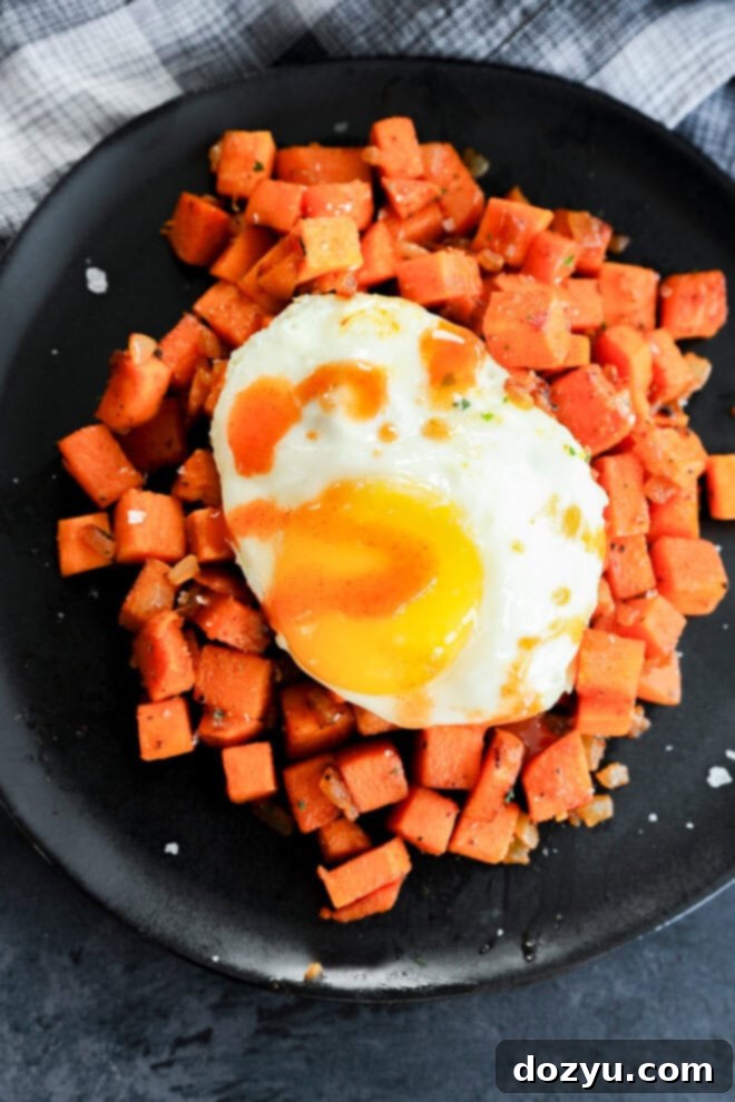 A black plate features a generous serving of smoky, diced sweet potatoes, perfectly golden and tender. It is crowned with a sunny-side-up egg, its bright yellow yolk shimmering, and garnished with a vibrant drizzle of hot sauce, suggesting a flavorful kick. The plate rests on a stylish black and white checkered cloth, enhancing the appetite appeal of this rustic breakfast dish.