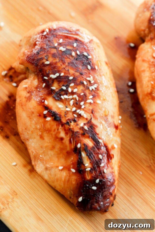 A grilled chicken breast, marinated in a savory Asian Chicken Marinade and topped with sesame seeds, is shown on a wooden cutting board. The chicken has visible grill marks and a glazed, slightly caramelized surface.