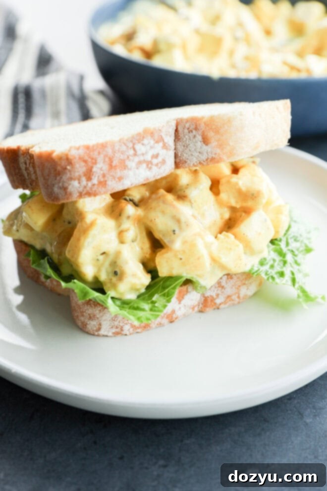 Golden Curried Chicken Salad 2 A close-up of an curried chicken salad sandwich with leafy lettuce on white bread, served on a white plate. In the background, a bowl of salad sits beside a dish of curried chicken salad for added variety.