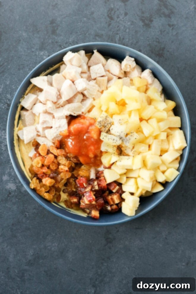 Golden Curried Chicken Salad 6 A bowl of curried chicken salad filled with chopped cooked chicken, diced pineapple, diced apples, golden raisins, chopped dates, chutney, and black pepper sits on a dark gray surface.