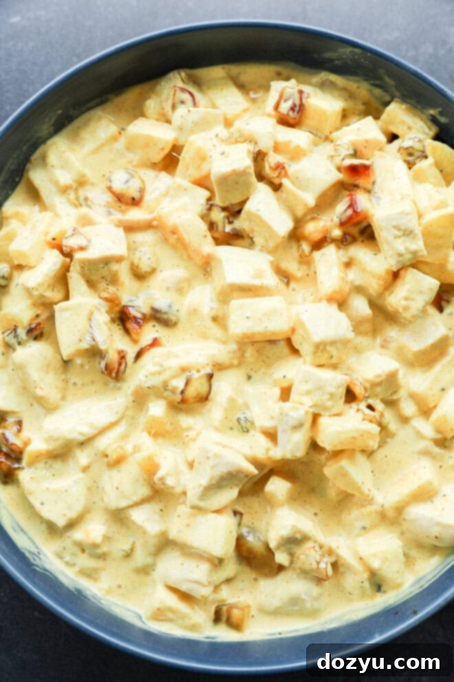 Golden Curried Chicken Salad 7 A close-up of a creamy yellow curry dish with cubed chicken, raisins, and fruit, all coated in a rich, spiced sauce for curried chicken salad, served in a dark bowl.