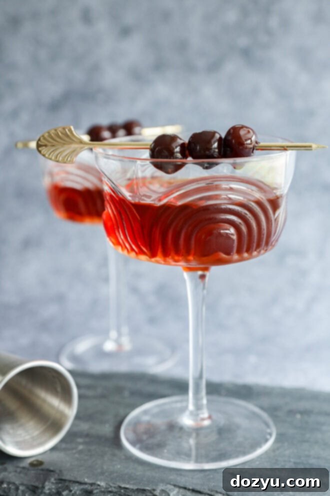 Crafting the Perfect Black Manhattan Cocktail 2 Two elegant cocktail glasses filled with a red Black Manhattan, each garnished with three dark cherries on a gold skewer. The wavy-patterned glasses rest on a slate surface, with a cocktail shaker nearby.