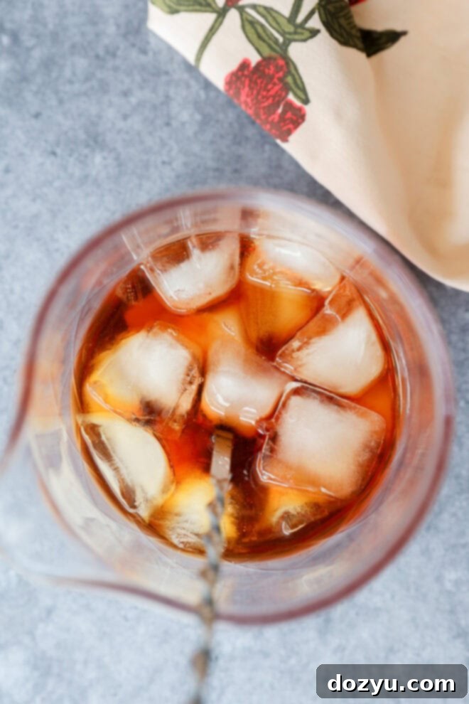 Crafting the Perfect Black Manhattan Cocktail 3 Overhead view of a glass filled with amber liquid and several ice cubes, likely a Black Manhattan, with a metal mixing spoon inside. A beige napkin with a red floral pattern is partially visible in the corner.