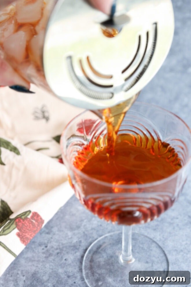 Crafting the Perfect Black Manhattan Cocktail 4 A Black Manhattan is being poured through a strainer into an elegant glass, with a floral-patterned napkin nearby on a gray surface.