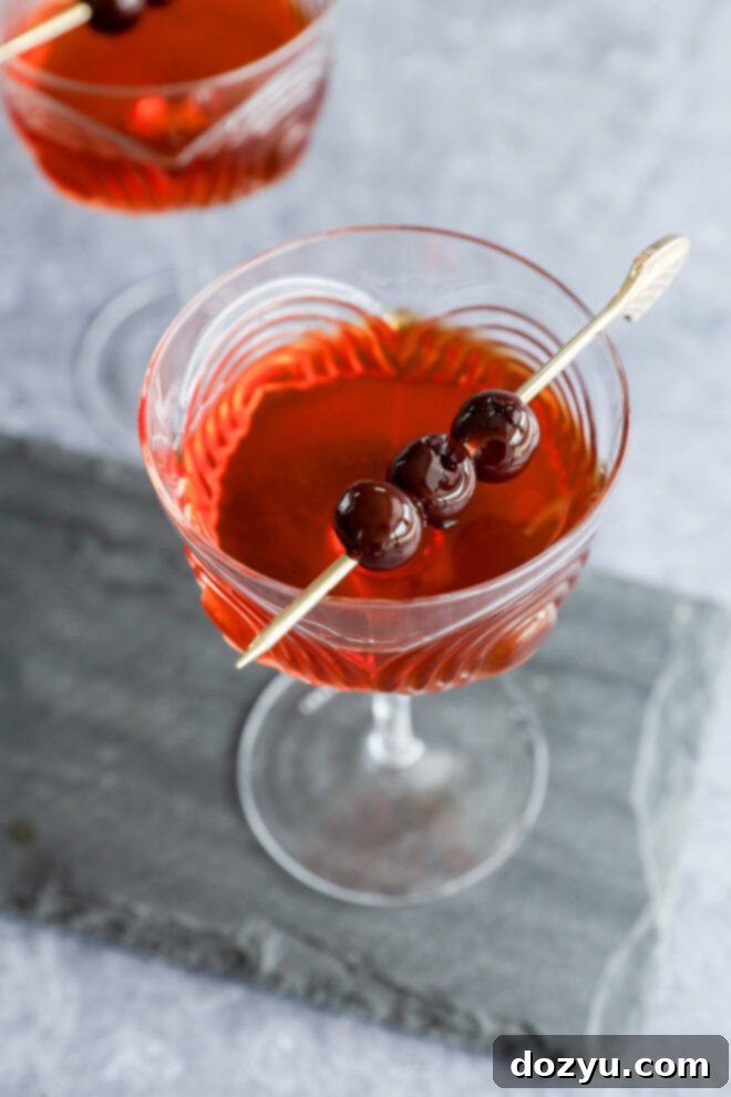 Crafting the Perfect Black Manhattan Cocktail 5 A Black Manhattan cocktail in a clear glass, garnished with three dark cherries on a cocktail pick, rests on a gray stone slab, with another similar drink partially visible in the background.
