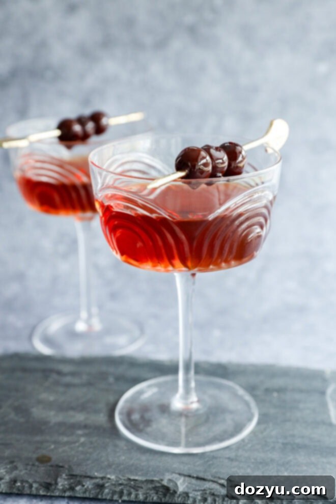 Crafting the Perfect Black Manhattan Cocktail 6 Two elegant coupe glasses filled with a Black Manhattan cocktail, each garnished with three dark cherries on a cocktail pick, set on a slate surface with a gray background.