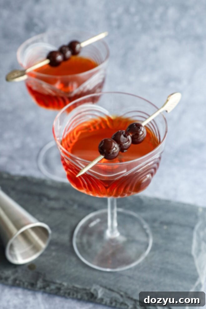Crafting the Perfect Black Manhattan Cocktail 7 Two crystal cocktail glasses filled with a reddish-brown Black Manhattan, each garnished with three dark cherries on a cocktail pick. A metal jigger sits nearby on a gray slate surface.