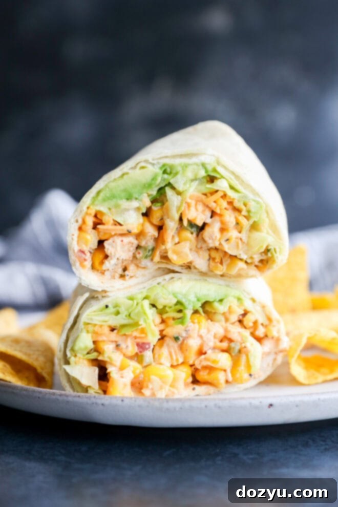 A close-up of a wrap cut in half, showcasing layers of crisp lettuce, vibrant corn, finely diced vegetables, and a rich, creamy chicken salad filling, elegantly presented on a plate next to a pile of crunchy tortilla chips.