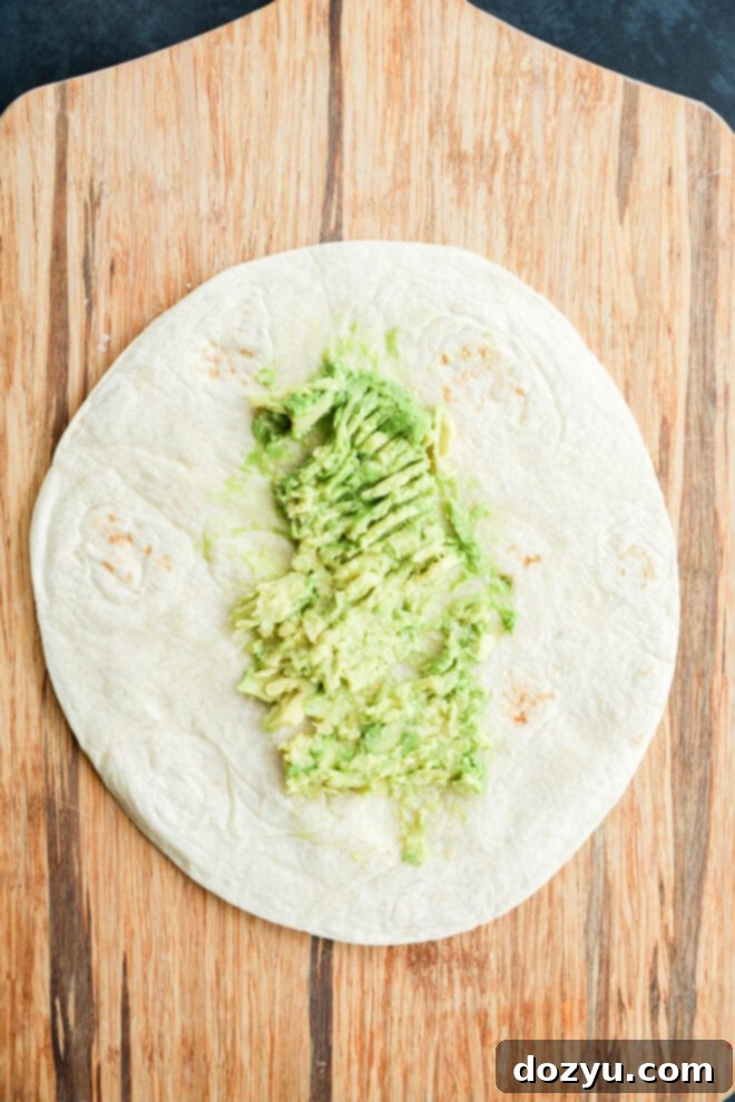 A flour tortilla resting on a wooden cutting board, with creamy, green smashed avocado spread evenly in the center, ready for additional toppings.