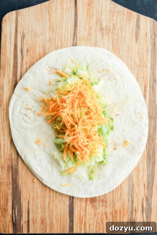 A flour tortilla on a wooden cutting board is neatly topped with a generous layer of shredded lettuce and shredded cheddar cheese, perfectly positioned and ready to be rolled into a delicious wrap.