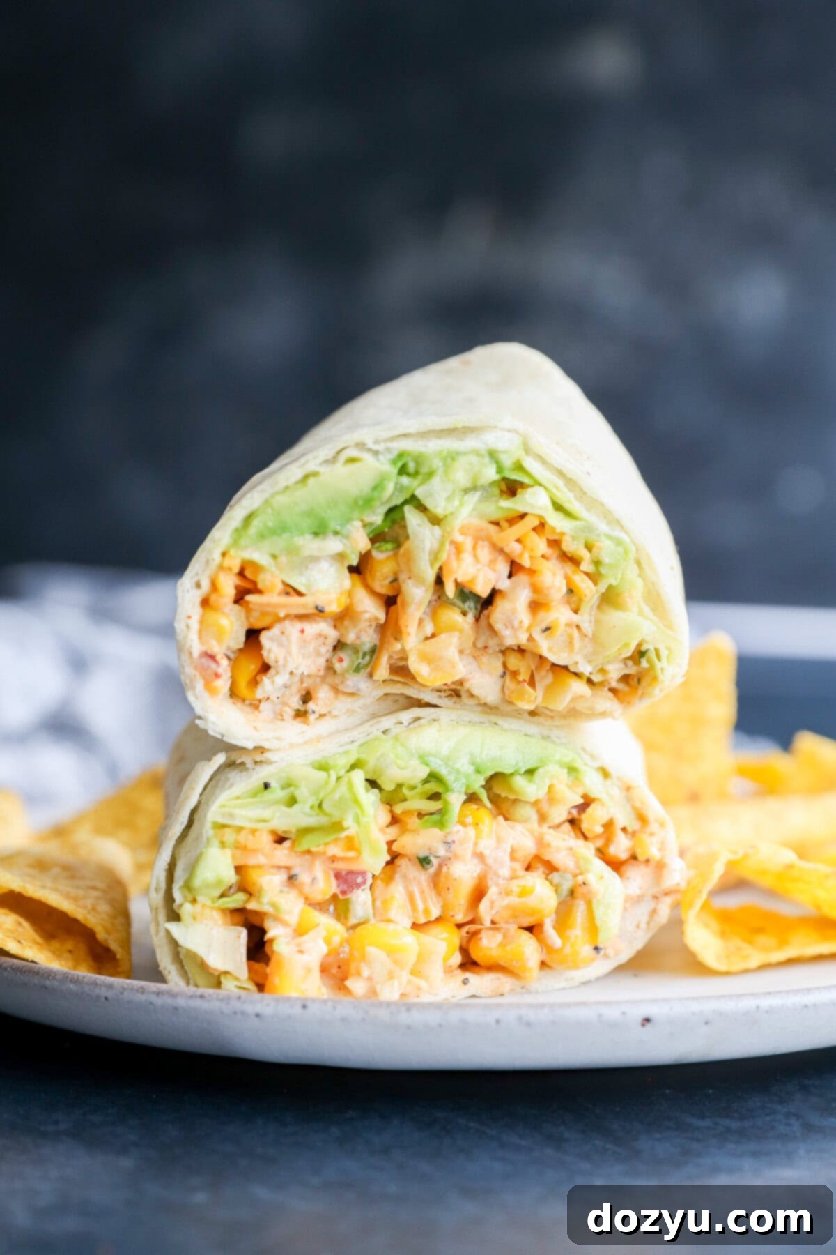 Two halves of a wrap filled with diced chicken, lettuce, and sauce are stacked on a plate with tortilla chips on the side, against a dark blurred background.