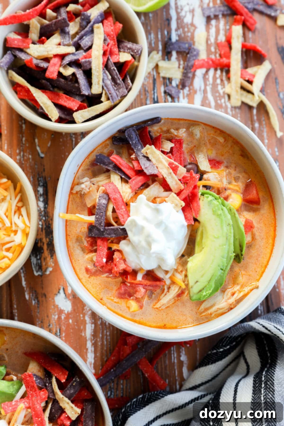 A bowl of stew topped with sliced avocado, sour cream, and colorful tortilla strips sits on a rustic wooden table, surrounded by bowls of shredded cheese and extra tortilla strips for the perfect finishing touch of this creamy chicken tortilla soup.