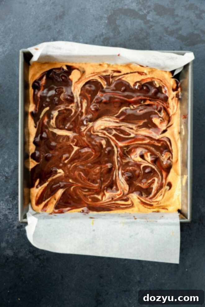 Spiced Pumpkin Fudge Brownies 6 A square pan lined with parchment paper holds a freshly baked dessert with a swirled chocolate and light batter pattern on top, resembling brownies or blondies. The background is a dark, textured surface.