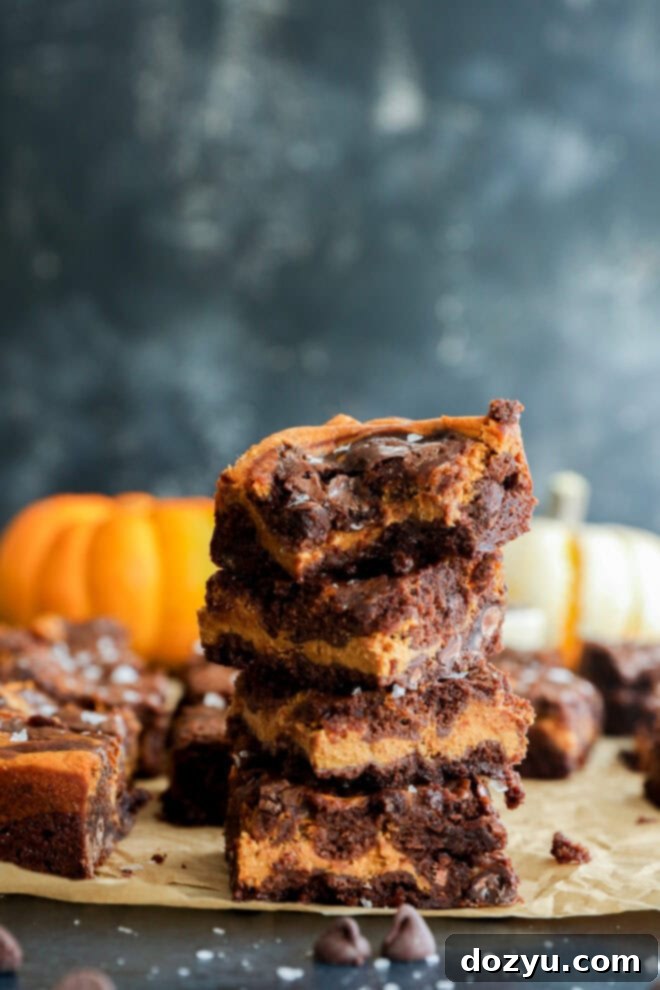 Spiced Pumpkin Fudge Brownies 9 A stack of four pumpkin chocolate chip brownies sits on parchment paper, with a bite taken out of the top brownie. In the background, there are pumpkins and more brownies, set against a dark, blurred backdrop.