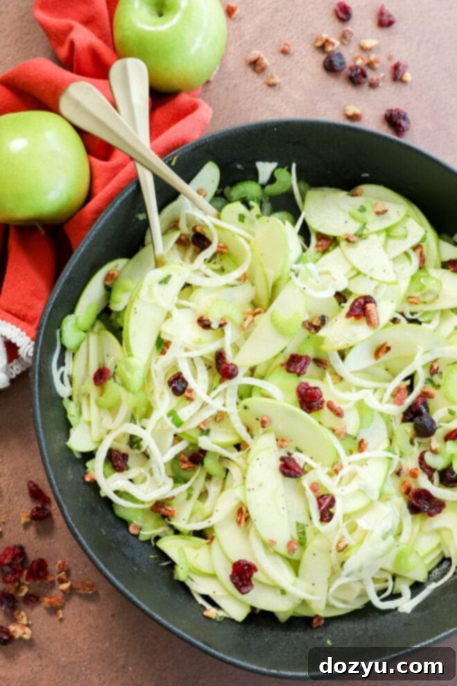 A bowl of fresh Apple Fennel Salad made with thinly sliced green apples, shredded cabbage, celery, dried cranberries, and chopped nuts. Two serving utensils rest in the bowl, with whole green apples and a red napkin beside it.