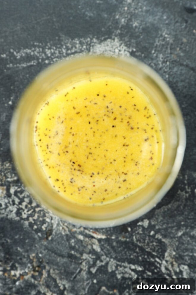 A close-up, top-down view of a glass jar filled with yellow salad dressing speckled with black pepper, set on a dark textured surface—perfect for drizzling.