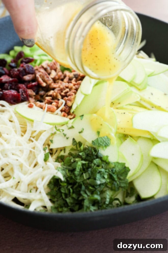 A close-up of sliced green apples, shredded fennel, herbs, dried cranberries, and nuts in a bowl, as a hand pours vinaigrette dressing from a jar over the ingredients.