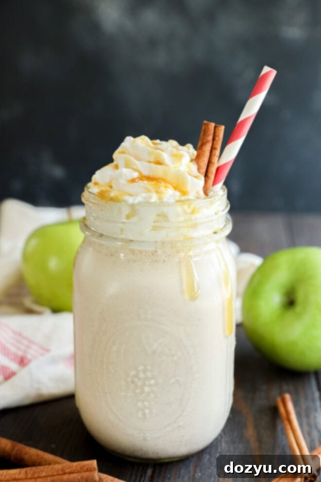 Velvet Apple Shake 6 A mason jar filled with a creamy milkshake, topped with whipped cream, caramel drizzle, two cinnamon sticks, and a red-striped straw. Green apples and cinnamon sticks are in the background on a dark wooden surface.