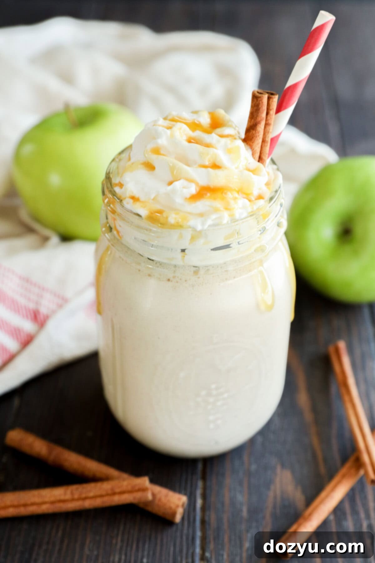 A creamy smoothie topped with whipped cream and caramel sauce in a mason jar, garnished with cinnamon sticks and a red-striped straw. Green apples and more cinnamon sticks are on the wooden table.