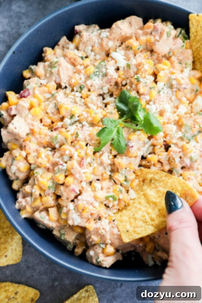 A bowl of creamy Mexican street corn chicken salad, topped with fresh cilantro and crumbled cheese, with a hand reaching in to scoop a generous amount onto a tortilla chip.