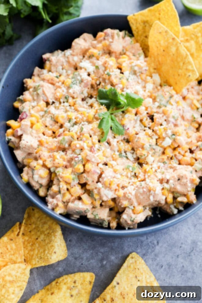 A blue ceramic bowl filled with creamy Mexican street corn chicken salad, garnished with crumbled Cotija cheese and fresh cilantro, surrounded by a scattering of tortilla chips on a textured gray surface.