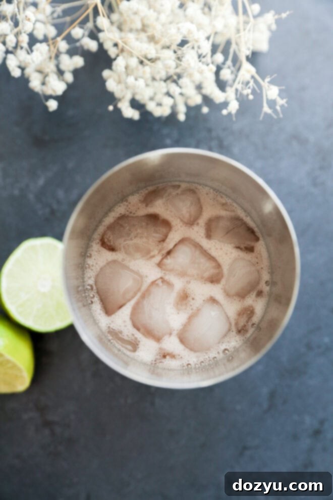 A metal cup filled with ice and a fizzy Prickly Pear Margarita sits on a dark surface, next to two lime halves and white dried flowers.