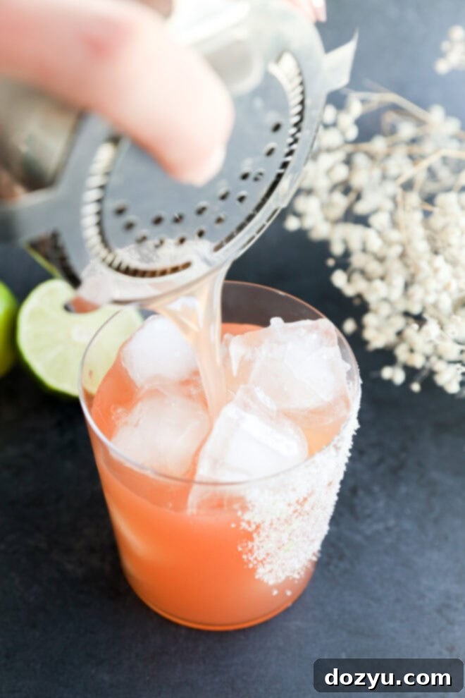 A hand pours a vibrant Prickly Pear Margarita over ice into a salt-rimmed glass, surrounded by lime slices and white flowers on a dark surface.