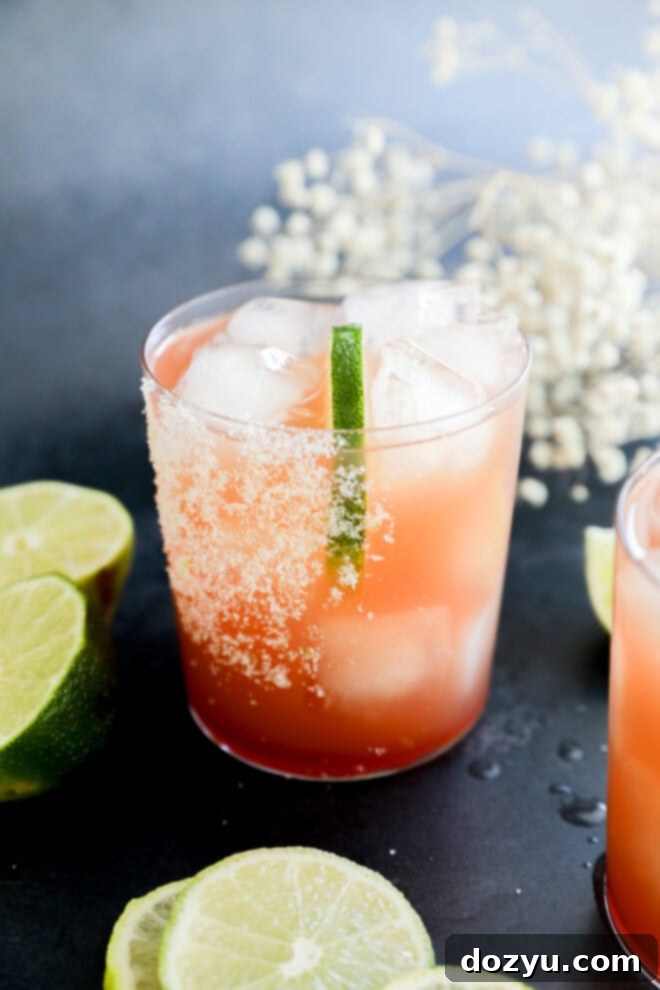 A glass filled with a vibrant Prickly Pear Margarita and ice cubes, garnished with a lime wedge and a salted rim. Slices of lime are scattered on the dark surface around the glass, with white flowers blurred in the background.