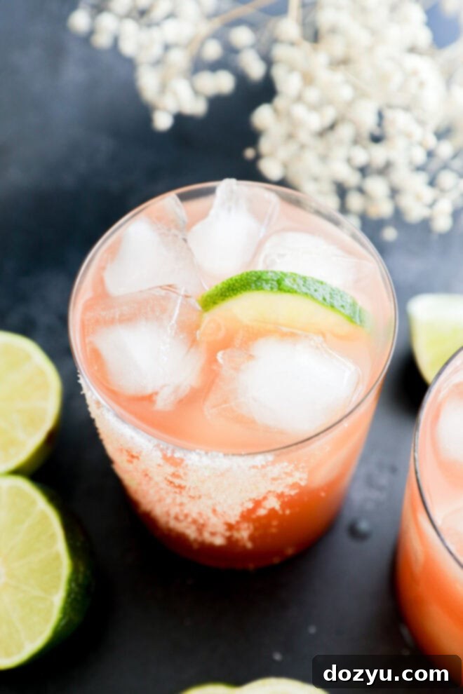 A glass filled with a pinkish-orange drink over ice, garnished with a lime wedge and a salted rim. Slices of lime and white flowers are visible in the background on a dark surface.