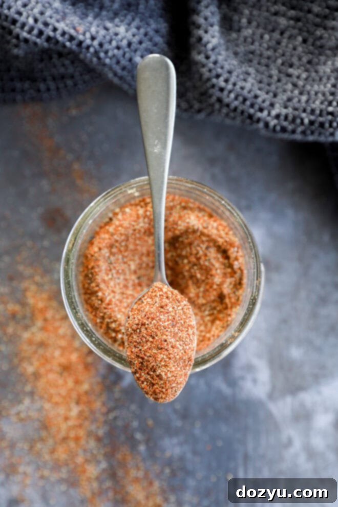 A spoon rests on top of a jar filled with a reddish-brown fajita seasoning blend, with some of the spice spilled on the dark surface below. A textured dark cloth is partially visible in the background.