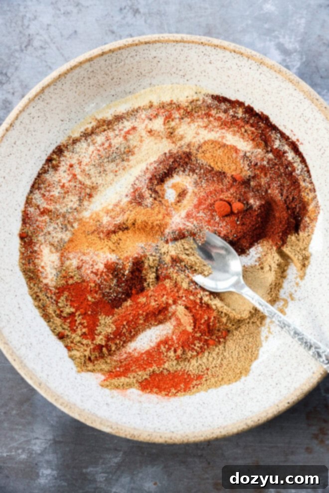 A white speckled bowl filled with various colorful ground spices being mixed together with a metal spoon on a gray surface.