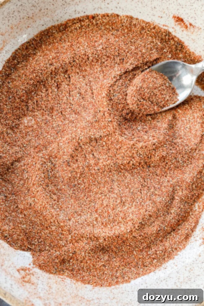 A close-up of a bowl filled with a reddish-brown spice blend, with a metal spoon resting in the mixture and some spices heaped on the spoon.
