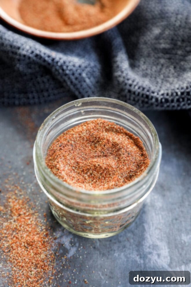 A small glass jar filled with a reddish-brown spice blend sits on a dark surface, with some of the spice spilled nearby and a textured dark cloth and a bowl in the background.