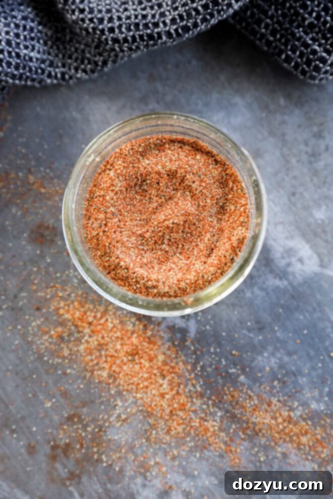 A glass jar filled with a reddish-brown spice blend sits on a gray surface, with some of the fajita seasoning spice scattered around the jar. A textured dark gray cloth is partially visible in the background.
