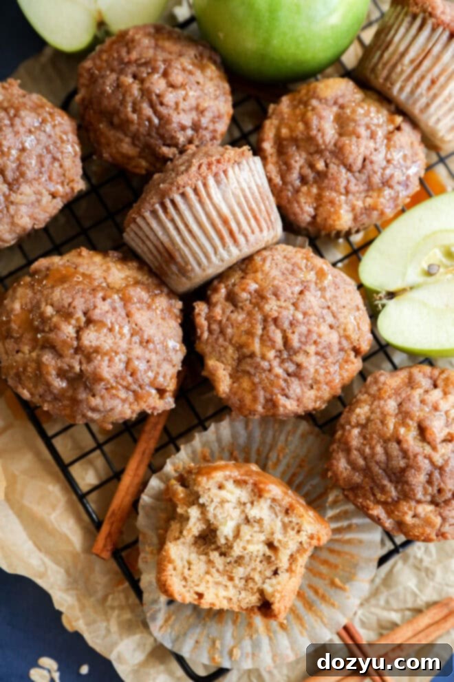 A wire rack holds several apple oatmeal muffins, some with paper liners. One muffin is cut open, showing a moist interior. Sliced green apples and cinnamon sticks rest on crumpled parchment paper nearby.