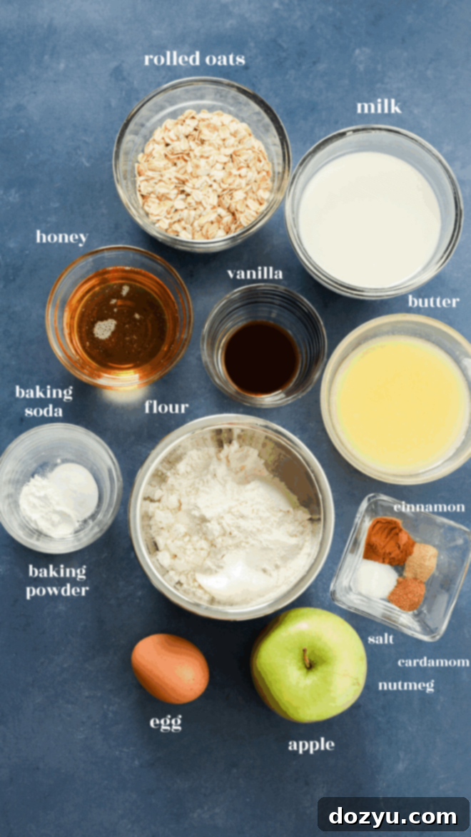 A flat lay of baking ingredients for apple oatmeal muffins on a blue surface, including rolled oats, milk, butter, vanilla, honey, flour, baking soda, cinnamon, spices, salt, an egg, and a green apple.