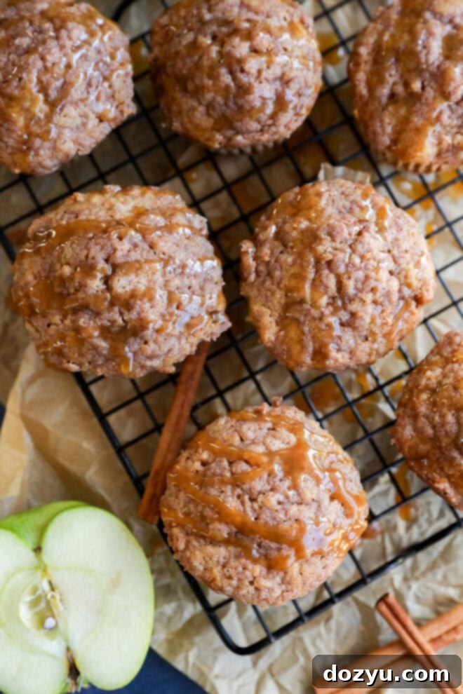 Apple oatmeal muffins topped with crumbly streusel and caramel drizzle are cooling on a wire rack. Cinnamon sticks and a sliced green apple rest nearby on parchment paper, completing this cozy scene.