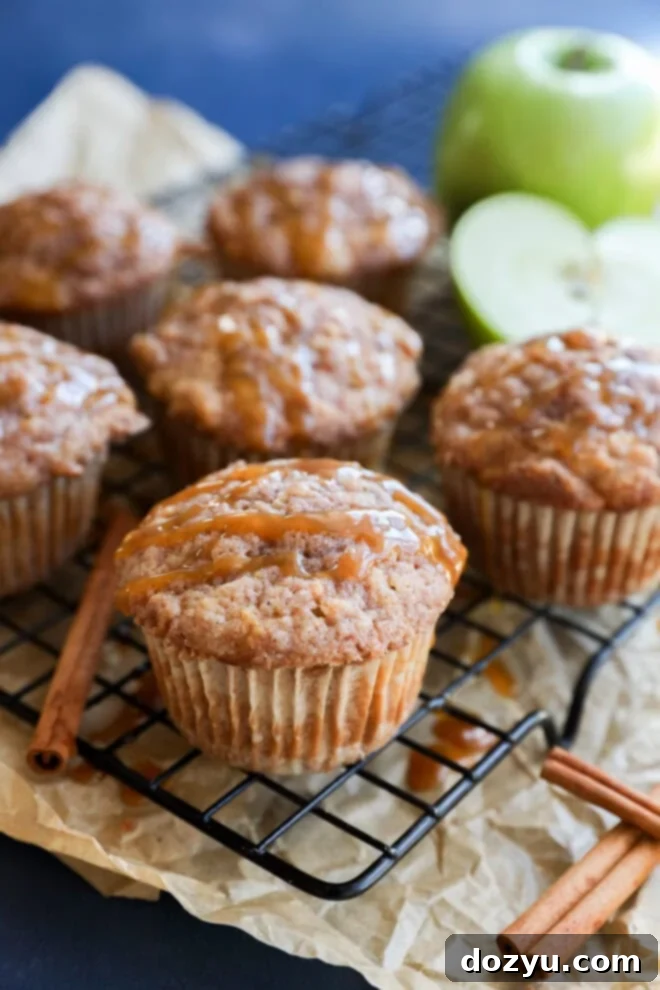 Freshly baked apple oatmeal muffins, drizzled with caramel, sit on a cooling rack lined with parchment paper. Cinnamon sticks and a green apple, both whole and halved, are visible in the background.