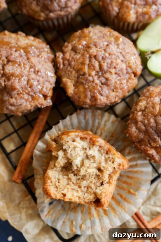 A close-up of apple oatmeal muffins on a wire rack, one muffin partially unwrapped with a bite taken, revealing a moist, fluffy interior. Cinnamon sticks and apple slices are nearby.
