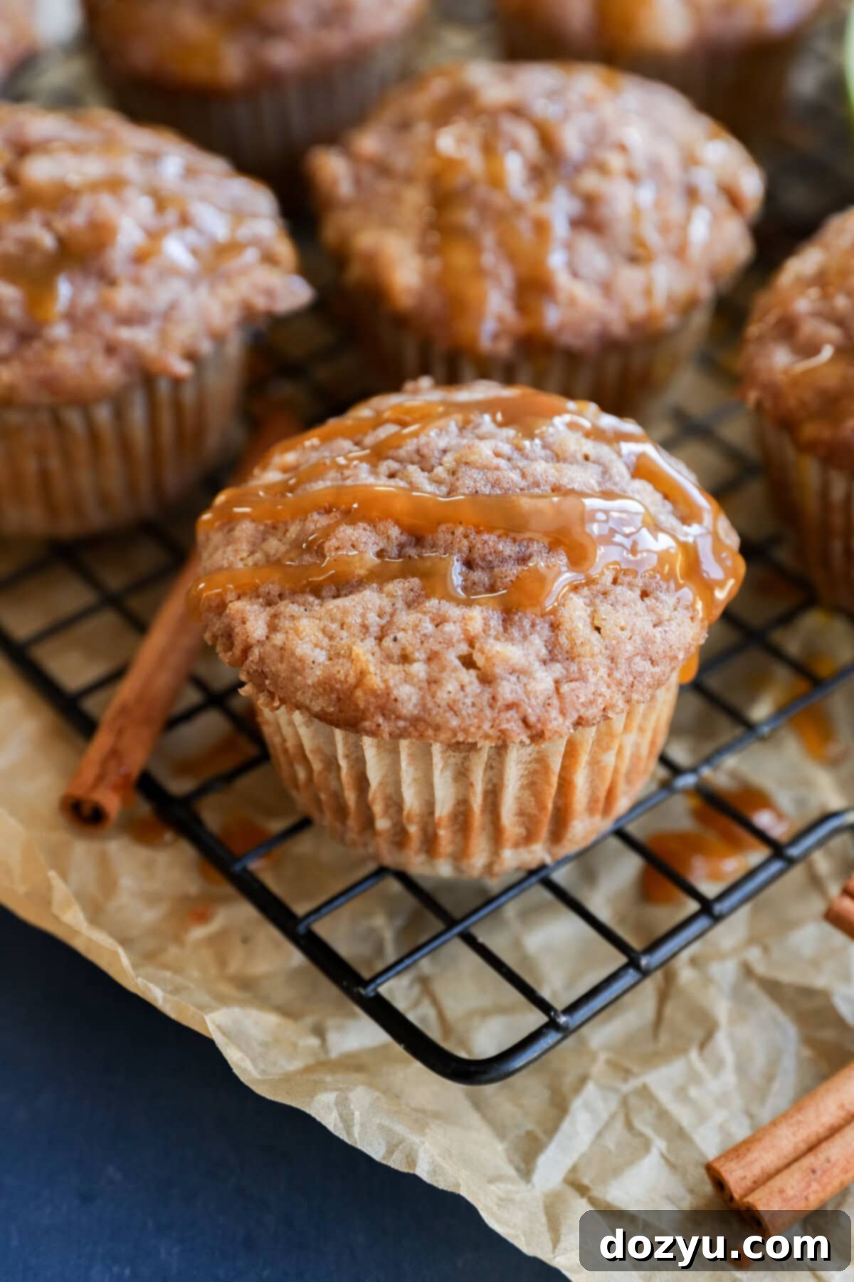 A close-up of cinnamon and apple oatmeal muffins drizzled with caramel sauce on a wire rack, with parchment paper and cinnamon sticks nearby.