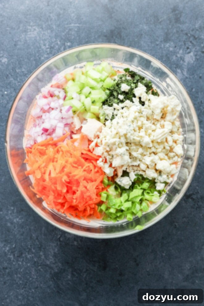 A glass bowl filled with onion, celery, shredded carrot, green onion, parsley, and crumbled cheese, all arranged in separate sections on top of a creamy mixture.