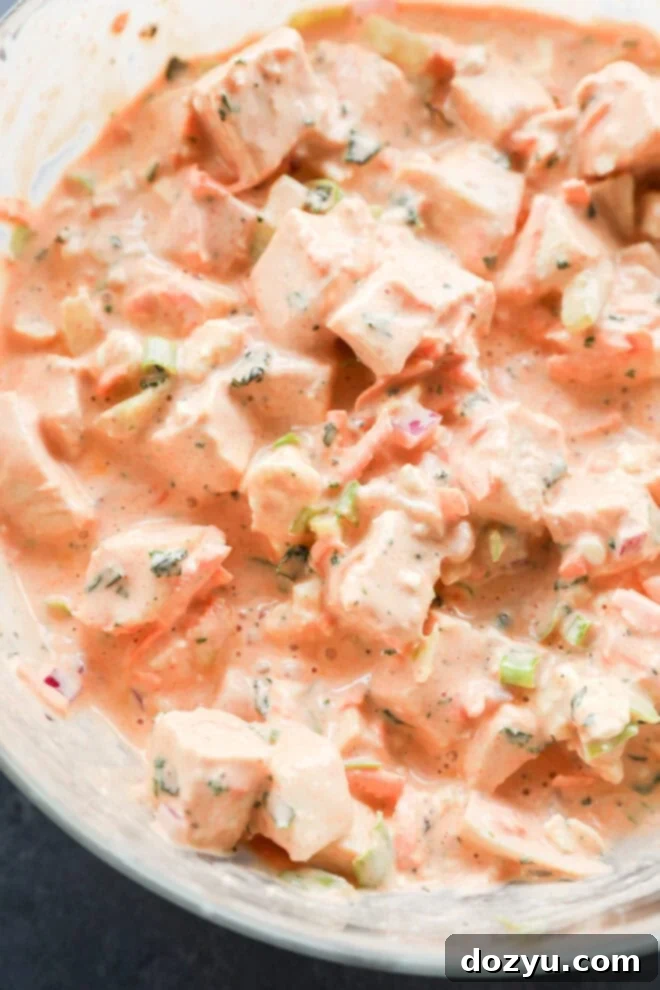 Close-up of a creamy, orange-tinted salad with chunks of chicken, diced vegetables, and chopped herbs mixed together in a bowl.
