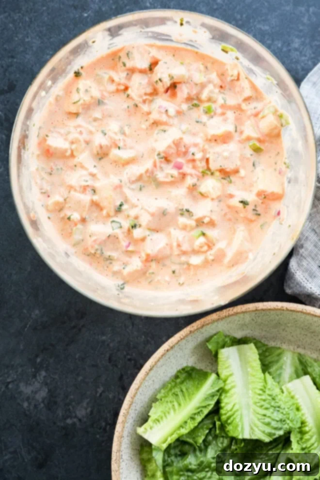 A glass bowl filled with a creamy mixture of chopped vegetables and herbs next to a ceramic bowl containing fresh romaine lettuce leaves, both on a dark tabletop.