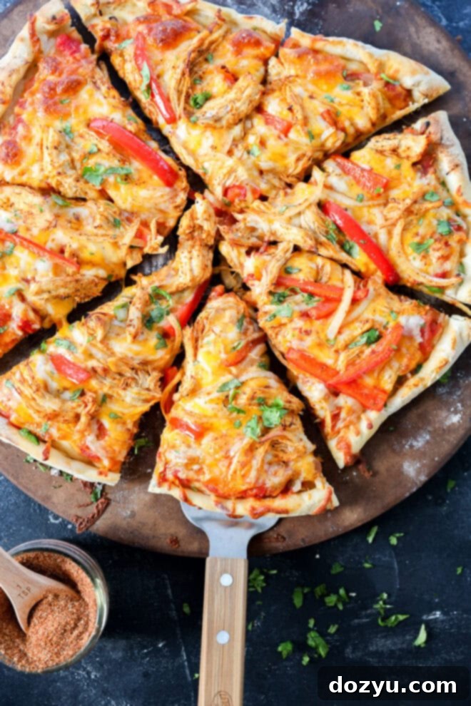Sizzling Fajita Pizza 2 A fresh, homemade pizza topped with shredded chicken, red bell peppers, melted cheese, and herbs sits on a round pizza stone. One slice is being lifted with a wooden-handled spatula. A small bowl of seasoning is nearby.